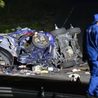 An investigator walks past a car on Thursday that crashed into a bridge in the village of Asuka, Nara Prefecture. Five students, all aged 19, died in the accident. The victims were found lying on the road, apparently after being thrown from the car. | KYODO