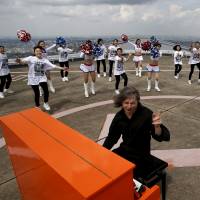 German musician Stefan Aaron plays the piano as cheerleaders perform next to him on top of the 296-meter-high Yokohama Landmark Tower, the second-tallest building in Japan, as part of his Orange Piano Tour, in Yokohama on Friday. | REUTERS