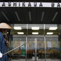 A Buddhist monk walks past Ashikaga Bank\'s head office in Utsunomiya in Tochigi Prefecture. Ashikaga Holdings Co., the bank\'s parent, is reportedly considering merging with Joyo Bank Ltd. | BLOOMBERG