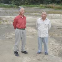 Kawamata Skating Club Chairman Toichi Kanno (right), 78, and Vice Chairman Hidekazu Ouchi, 66, visit an idle rice paddy that will again host its well-known skating rink in Kawamata, Fukushima Prefecture, in January. | FUKUSHIMA MINPO