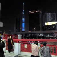 People take photos Saturday in Tokyo\'s Asakusa district from a vantage point where Sumida Ward\'s Tokyo Skytree could be seen illuminated in blue. To the right is Asahi Breweries Ltd. headquarters with its distinctive golden flame that sits atop the building. | YOSHIAKI MIURA