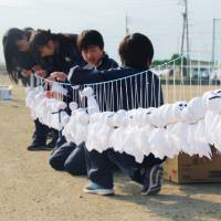 Students at Takase Middle School in the town of Ikeda, Nagano Prefecture, are seen next to <em>teruterubozu</em> dolls hanging on a line in the school playground in October. On Friday, the school announced that Guinness World Records has recognized it after students hung more than 10,000 dolls on a single line. The simple handmade cotton dolls are traditionally hung under house eaves to pray for good weather the following day. | KYODO
