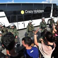 Migrants and refugees leave by bus as solidarity groups protest behind a line of riot police during the forced evacuation of a makeshift camp close to the Greece-Macedonia border near the village of Idomeni on Tuesday. Greek police on May 24 moved hundreds of migrants out of the overcrowded camp of Idomeni, launching a major operation to clear up the squalid tent city where thousands fleeing war and poverty had lived for months. | AFP-JIJI
