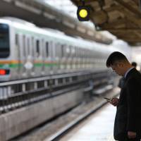 A man uses a mobile phone in front of an East Japan Railway Co. train at Tokyo Station. According to a recent survey by JR East, over 20 percent of train delays caused by objects falling onto tracks were attributable to dropped smartphones. | BLOOMBERG