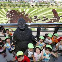 Children pose Wednesday near a paddy where rice has been planted in the shape of Godzilla. The huge image in the Aomori Prefecture village of Inakadate uses nine kinds of rice. It promotes the movie \"Shin Godzilla,\" which is due for release this month. | KYODO
