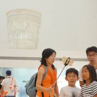 Visitors take selfies below a giant replica of Cup Noodles displayed at the Cup Noodles Museum in Yokohama on Aug. 15. | SHUSUKE MURAI