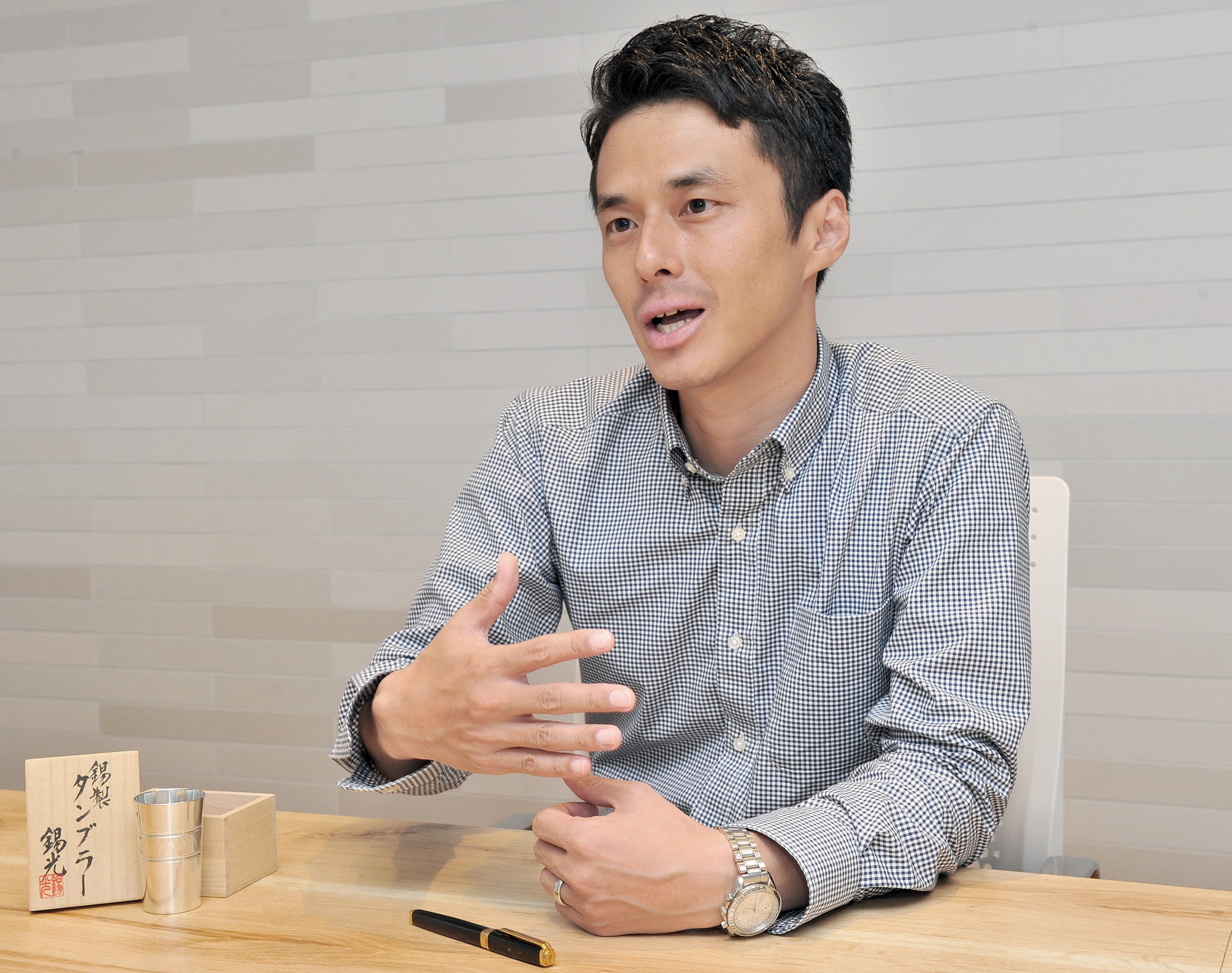 Keigo Omaki talks about his effort to document Japan's traditional craftsmen online, during an interview in Tokyo on Aug. 25. Before him is a pewter cup made by Keiichi Nakamura, one of the craftsmen featured in the video project. | YOSHIAKI MIURA
