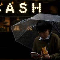 A Tokyo pedestrian juggles her umbrella, wallet and smartphone. With wages static, the Bank of Japan is targeting bond yields to try to reach its 2 percent inflation target. | REUTERS