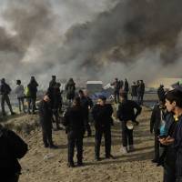 Migrants, police and media stand on a hill overlooking shelters burning in the makeshift migrant camp near Calais, northern France, Wednesday. French authorities declared the Calais migrant camp known as \"the Jungle,\" empty on Wednesday, after fires set by departing migrants accelerated plans to evacuate the burgeoning slum. | AP