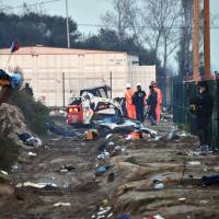 Police officers with masks covering their face stand guard as members of the demolition crew watch a mechanical digger tearing down the \"Jungle\" migrant camp in Calais, northern France, on Wednesday. | AFP-JIJI
