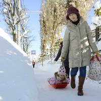 A young girl smiles as she is pulled on a sledge along a snow-covered sidewalk in Higashi Ward, Sapporo, on Saturday afternoon. | KYODO