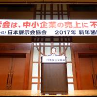 Japan Exhibition Association Chairman Tadao Ishizumi speaks in front of a slogan that reads \"Exhibitions are essential for small and midsize firms\' sales\" at JEXA\'s New Year\'s party in Tokyo on Jan. 12. | SHUSUKE MURAI
