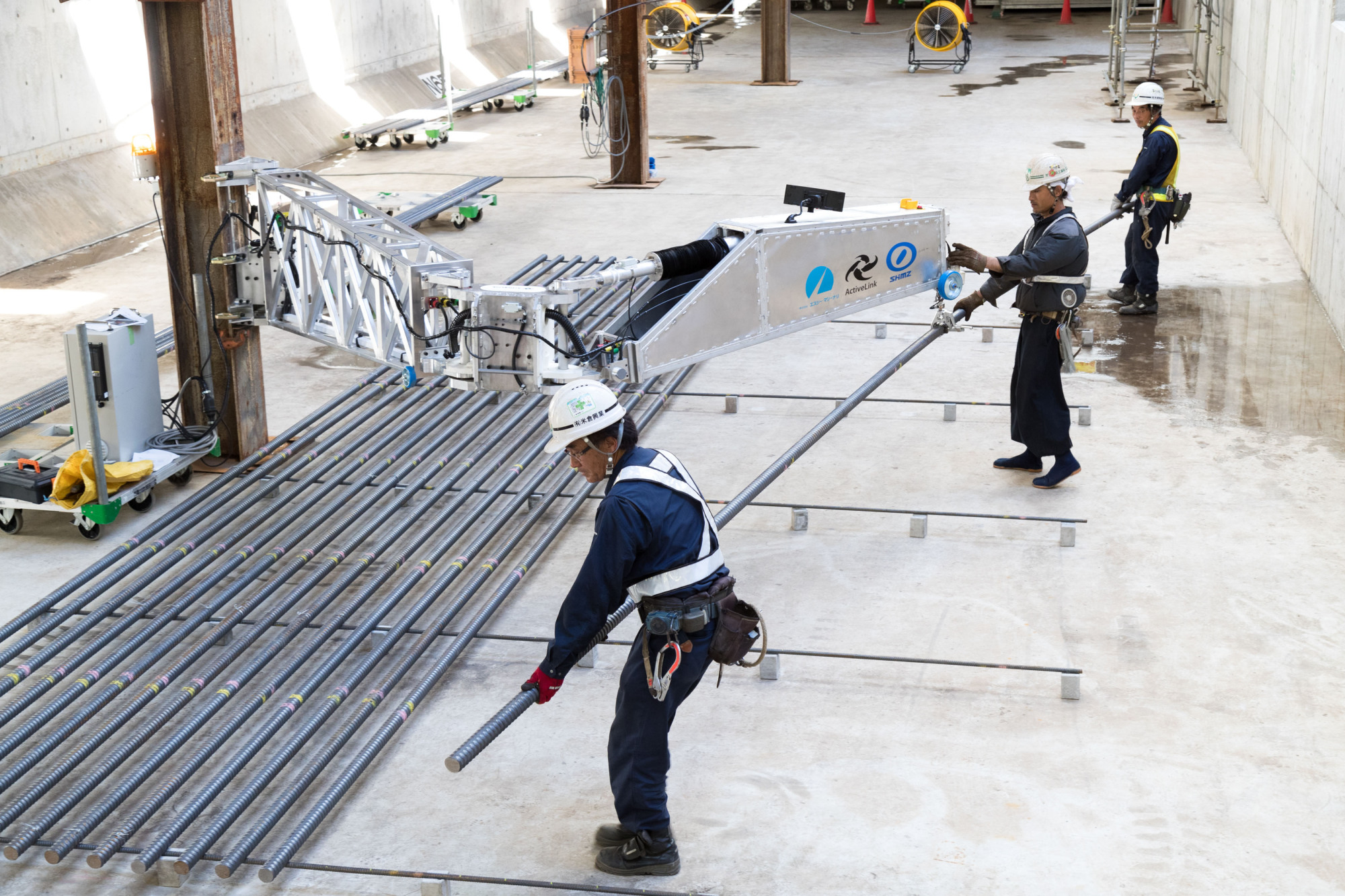 An arm-shaped robot developed by Shimizu Corp. helps construction workers lift 200-kg steel rods in Chiba Prefecture last August. | SHIMIZU CORP. / VIA KYODO