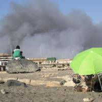 People sit on the beach as black smoke rises from wildfires at the nearby Castelfusano pine forest, near Rome Monday. | MASSIMO PERCOSSI / ANSA / VIA AP