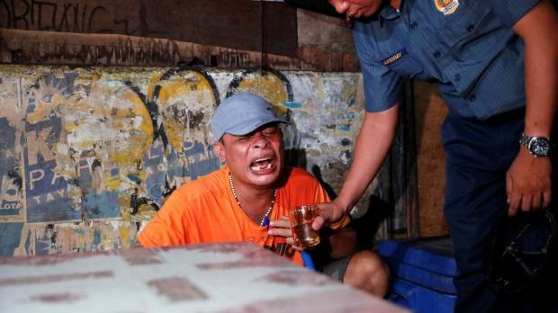 A police officer tries to comfort a man by giving him a glass of water after his brother was killed in a spate of drug-related violence Wednesday night. Philippine war on drugs and crime intensifies, with at least 58 killed in three days