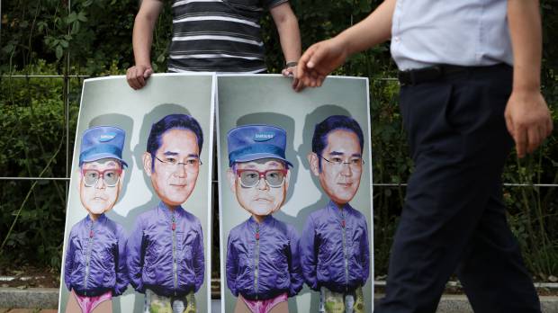 A protester shows images of Samsung heir Lee Jae-yon and his father, Lee Kun-hee (in hat), South Korea’s richest man, Friday in Seoul. Samsung heir Lee Jae-yong sentenced to five years in prison for bribery