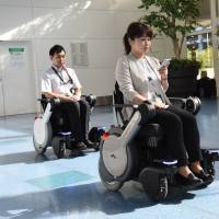 Officials at Tokyo\'s Haneda airport on Tuesday demonstrate self-navigating electric wheelchairs that will allow users to call one up with smartphones and use them to travel anywhere within the airport. With sensors and other safety devices built in, the wheelchairs can automatically avoid obstacles. Along with foreign tourist-friendly signs and other features, the wheelchairs will be available as early as next April. | SATOKO KAWASAKI