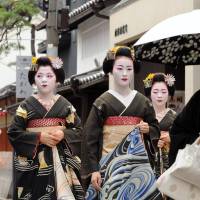 Apprentice geisha, known as maiko, clad in formal black kimono, walk along a street in Kyoto\'s Gion district on Tuesday. They were taking part in the annual summer hassaku, a customary event in which the traditional entertainers pay courtesy calls on their teachers and teahouses to express their gratitude. | KYODO