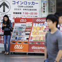 A woman uses a smartphone next to a display advertising products for KDDI Corp.’s au brand, NTT Docomo Inc. and SoftBank Group Corp. outside an electronics store in Tokyo in September 2015. What would a 40% cut to phone bills mean for Japan's mobile carriers?