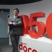 Hisakazu Tsuboya of NTT DOCOMO poses by signage for the company’s next-generation cellular network. In an interview, he discussed the first year of commercial 5G service in Japan and how it might develop in the future. |  NTT DOCOMO
