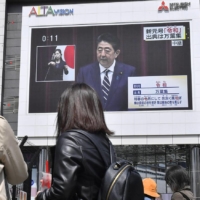 Left: Shoppers watch Abe explain the meaning of Japan’s new era name Reiwa on a giant screen in Tokyo’s Shinjuku Ward on April 1, 2019. | KYODO